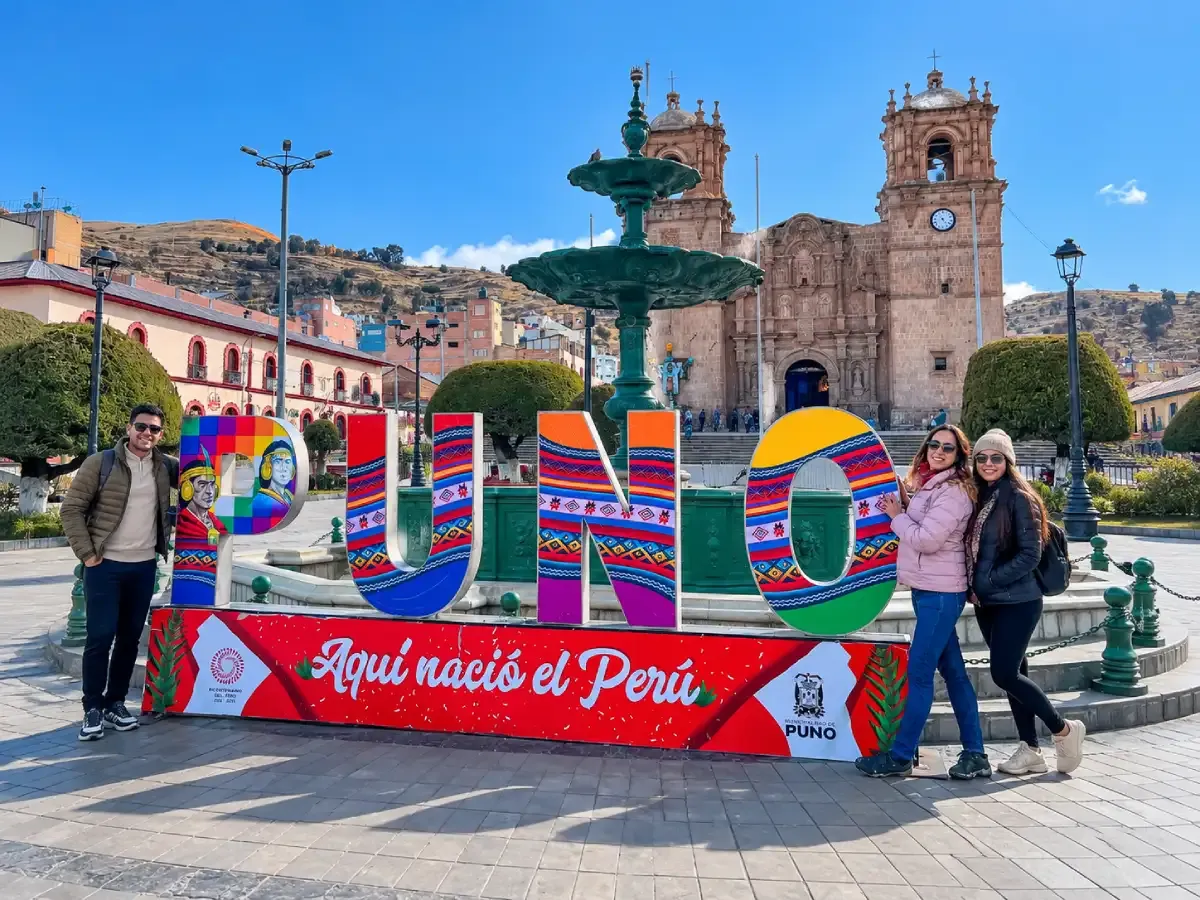 Tres turistas posando junto a las coloridas letras de Puno, con la Catedral de Puno y la fuente de la Plaza de Armas al fondo, durante un recorrido turístico por la ciudad de Puno, Perú.