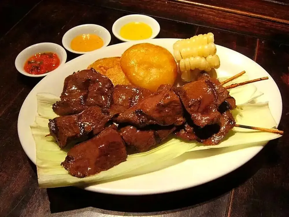Anticuchos de corazón con papa dorada, choclo y salsas en un plato blanco.