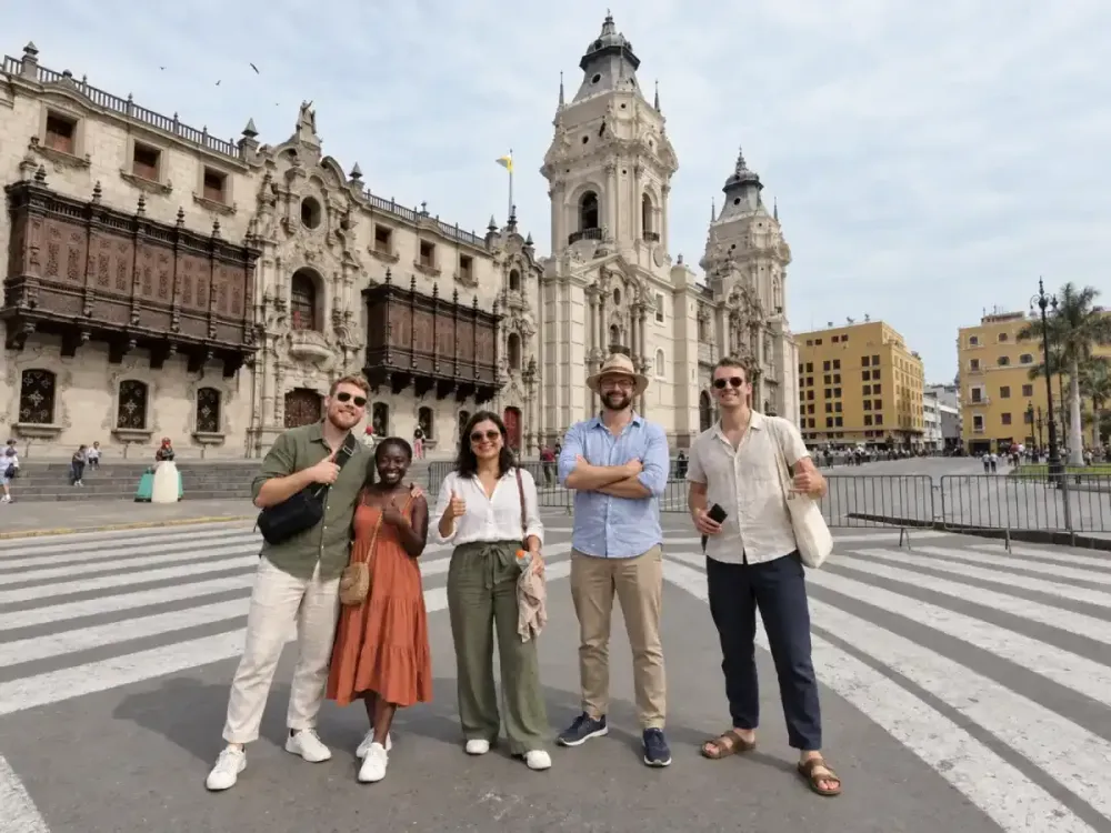 Un grupo de cinco turistas sonrientes posa en la Plaza Mayor de Lima, con la fachada de la Catedral de Lima y los tradicionales balcones coloniales del Palacio Arzobispal de fondo bajo un cielo despejado.
