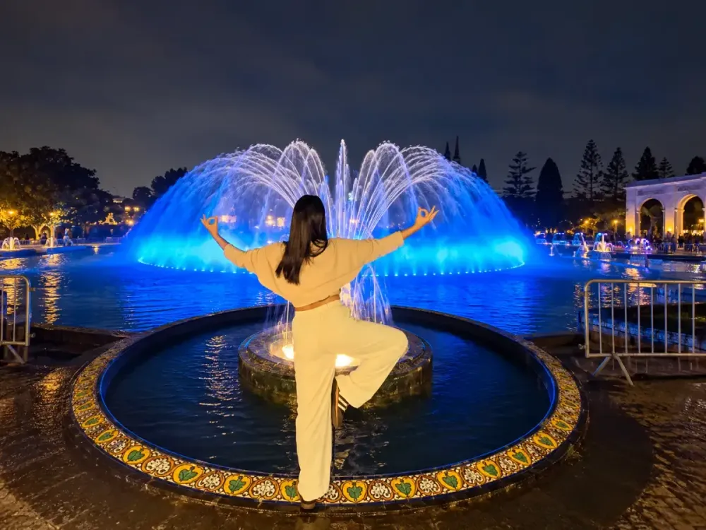 Mujer haciendo una postura de yoga de noche frente a la Fuente Mágica iluminada.