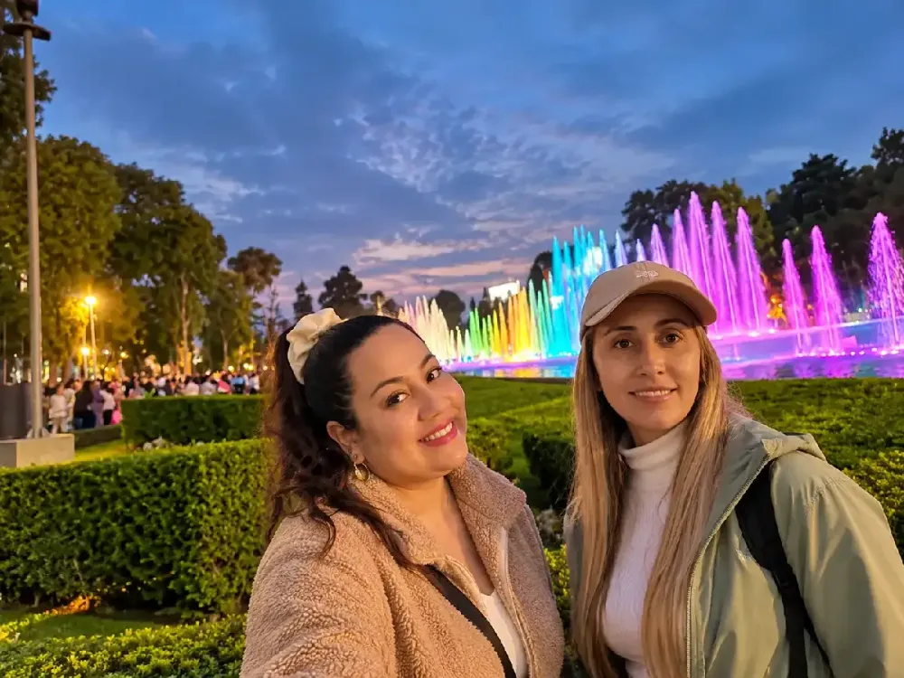 Dos amigas sonrientes posan de noche frente a la Fuente de la Fantasía iluminada.