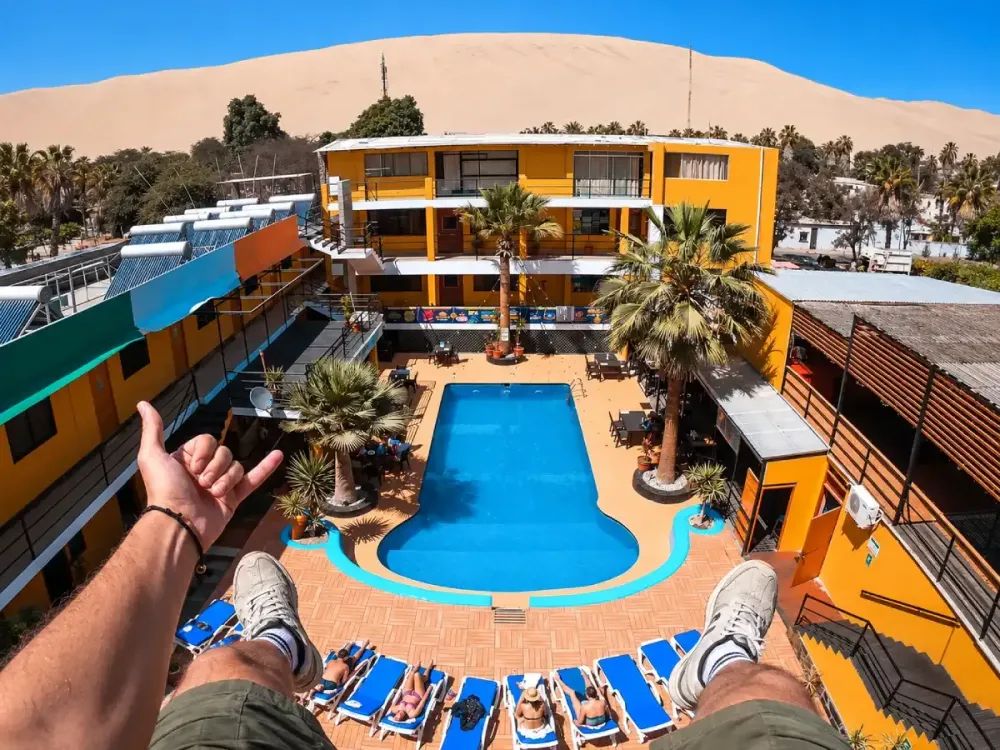 Vista de una piscina en un alojamiento de Huacachina, rodeada de palmeras y edificios, con una gran duna del desierto al fondo.
