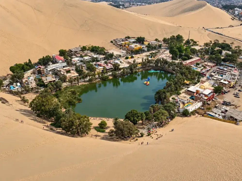 Vista aérea del oasis de Huacachina, rodeado de palmeras, alojamientos y grandes dunas del desierto de Ica, Perú.