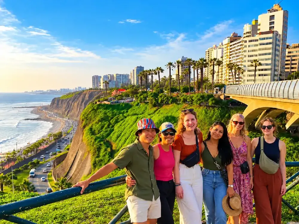Un grupo de jóvenes turistas posa sonriente junto a la baranda del Malecón de Miraflores. Al fondo se aprecian los acantilados verdes de la Costa Verde, el océano Pacífico, la autopista costera y los modernos edificios residenciales bajo un cielo despejado.