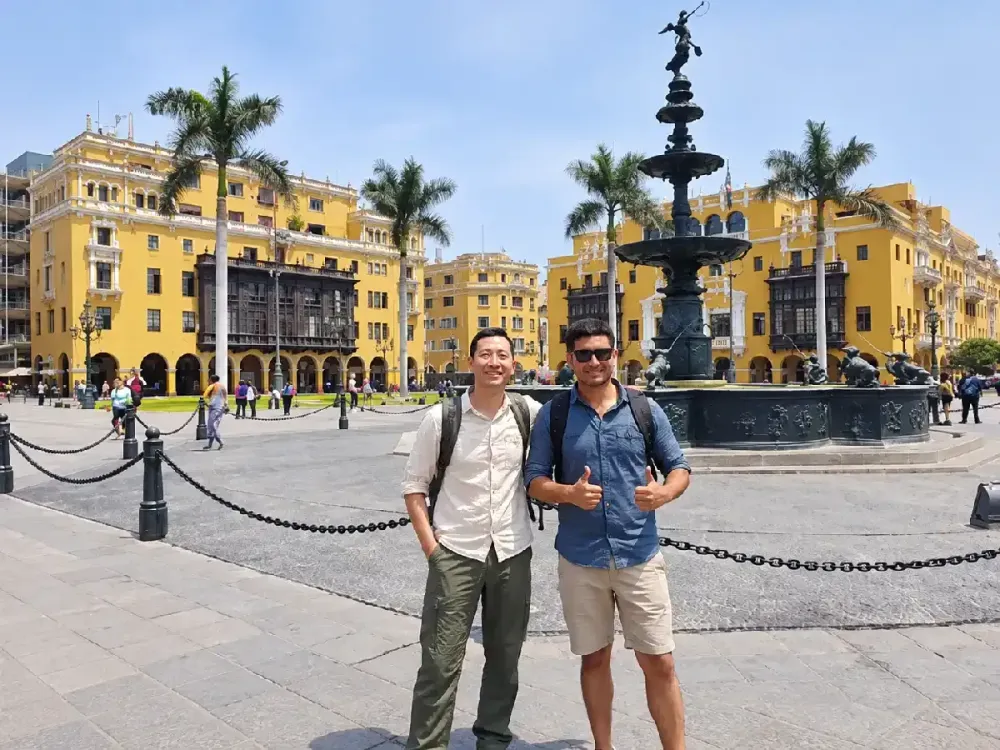 Dos amigos posan sonrientes junto a la pileta colonial de la Plaza Mayor de Lima.