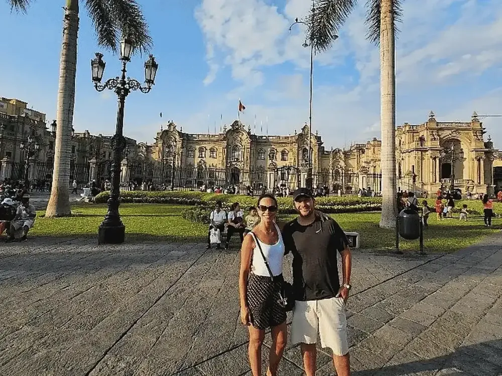 Hombre y mujer posando en el centro de la Plaza de Armas de Lima durante el día. De fondo destaca la arquitectura clásica del Palacio de Gobierno y transeúntes descansando en los jardines.