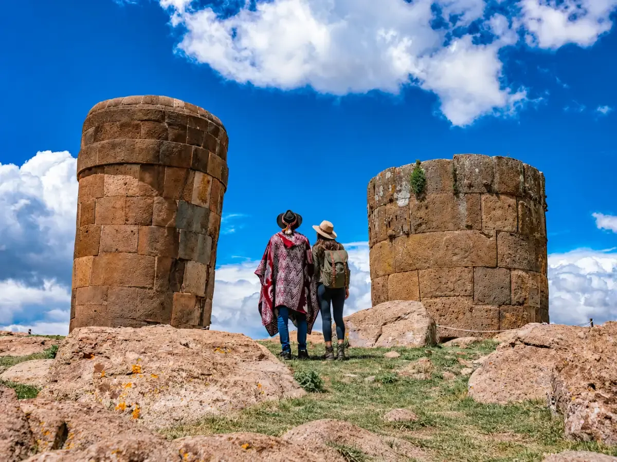 Dos turistas de espaldas contemplando las chullpas de Sillustani en Puno, Perú, rodeadas de un paisaje andino bajo un cielo azul intenso.