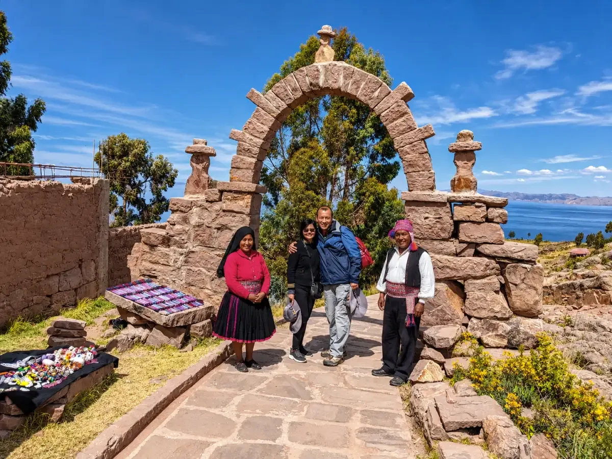 Pareja de turistas junto a dos pobladores con vestimenta tradicional en la isla de Taquile, con el lago Titicaca y un arco de piedra al fondo.
